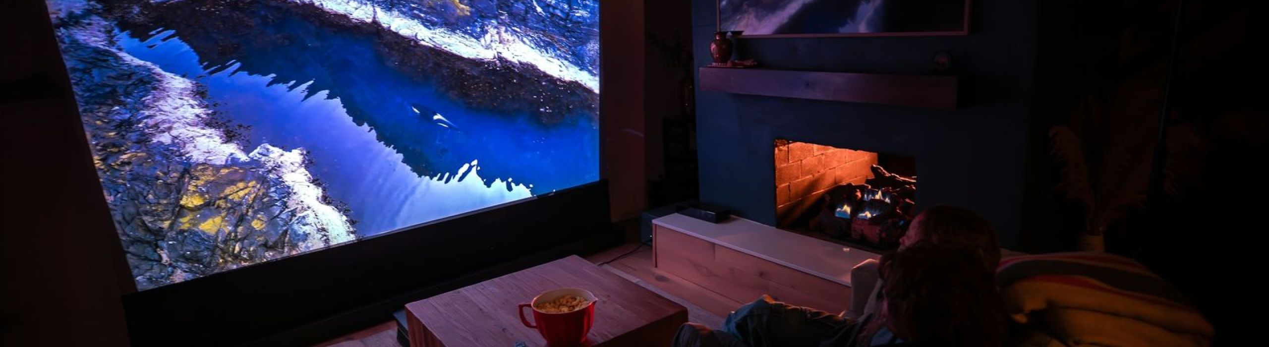 Living room at night lit only by a large projected image and a glowing fireplace, with a sofa facing the screen