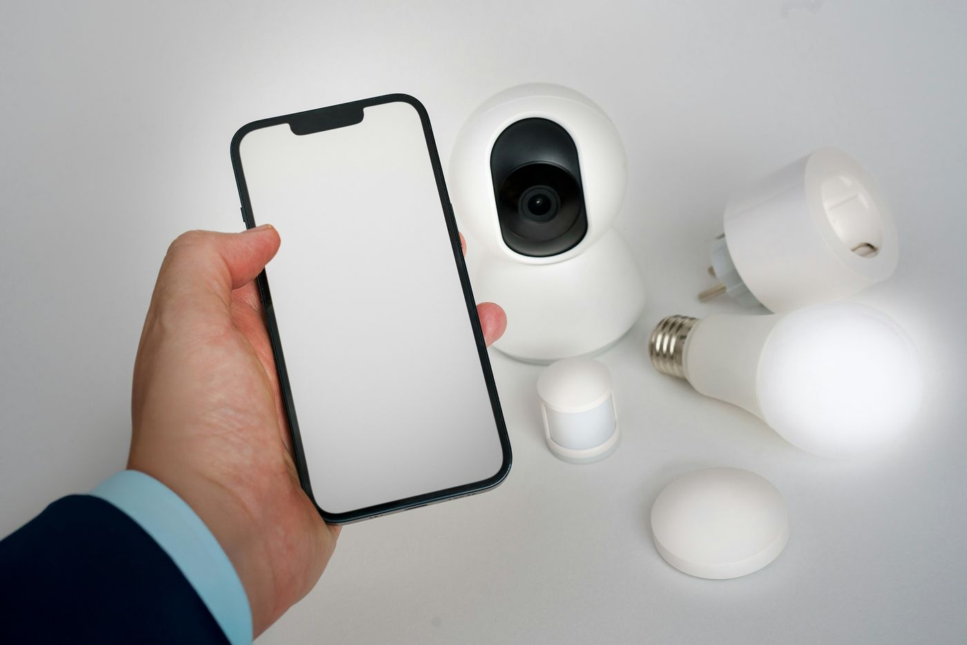 Hand holding a smartphone next to a home camera, smart plug and LED bulb