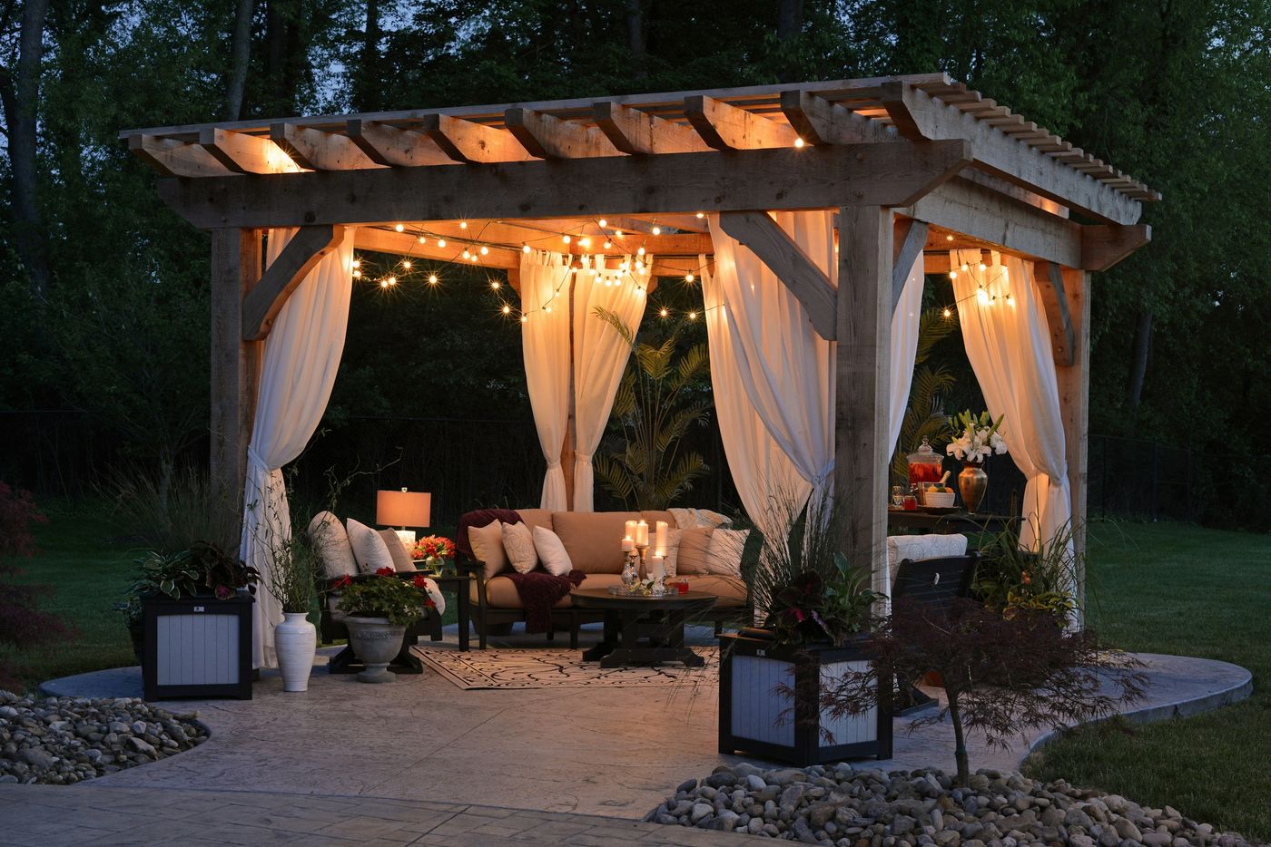 Backyard pergola with drapes, outdoor seating and string lights glowing at night